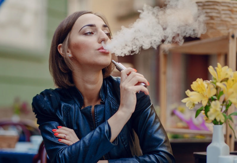 A woman wearing a brown fur coat exhales a thick cloud of vapor while vaping outdoors, with blurred trees in the background.
