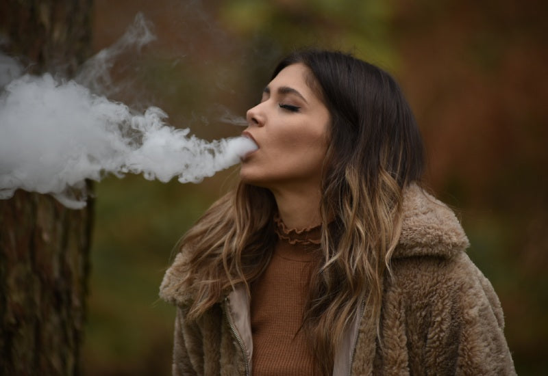 A woman outdoors exhaling a thick cloud of vapor while vaping, wearing a brown fur-lined coat and a brown top, with blurred trees in the background.